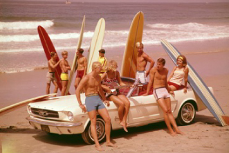 A group of surfers with their surfboards sitting and standing around a convertible on a beach