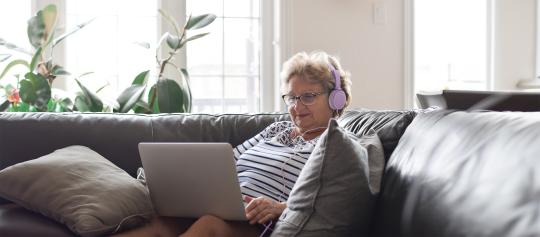 An old lady relaxing on a couch looking at a computer monitor with her headphones.