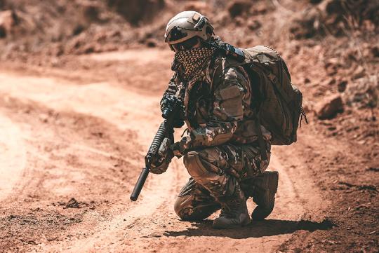 An army serviceman holding a rifle, dressed in combat gear in the middle of a dirt road